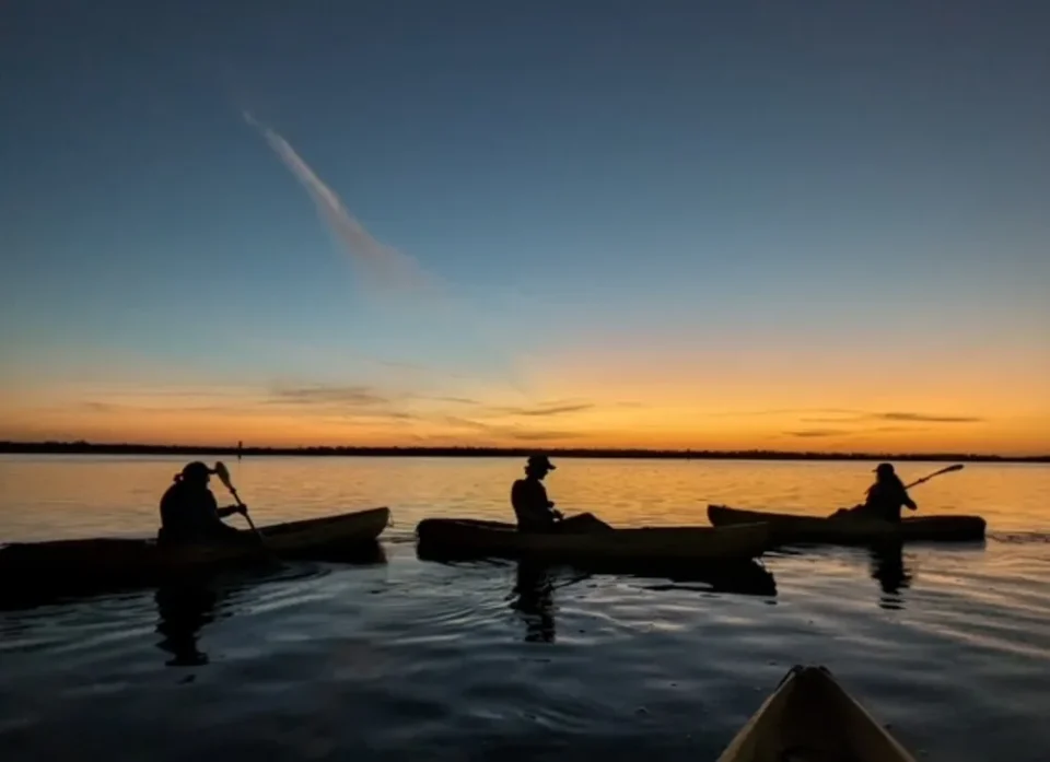 Silhouettes of three kayakers on the water at sunset in waters of Fort Myers, FL. A lovely gradient of blue and orange.